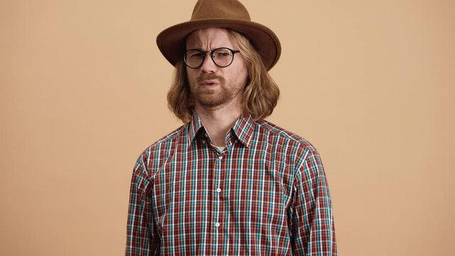 The man in the hat and eyeglasses saying no and wrinkling his face while standing in a beige studio