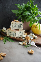 Gorgonzola on a wooden plate next to arugula and walnuts. Cheese with a noble blue mold on a gray table and
dark background.
