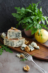 Gorgonzola on a wooden plate next to arugula and walnuts. Cheese with a noble blue mold on a gray table and
dark background.
