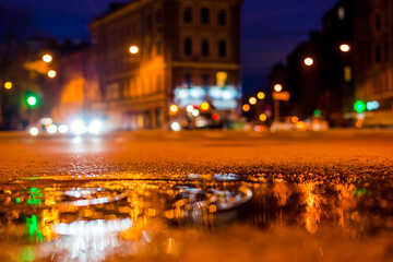Fototapeta na wymiar Nights lights of the big city, the night intersection between the houses on which cars travel. Close up view of a puddle on the level of the hatch