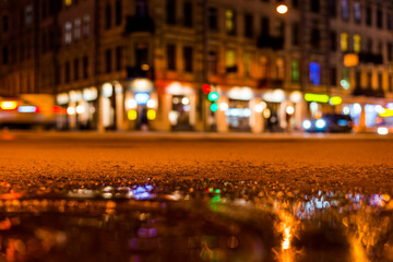 Nights lights of the big city, the night street with driving cars. Close up view of a puddle on the level of the hatch