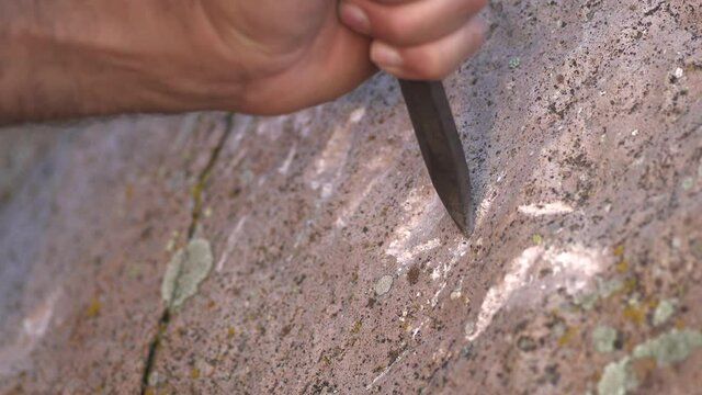 Prehistoric petroglyph carving reenactment using iron chisel on stone surface, close detail. Primitive rock inscription scene shows hand tools etching symbols into boulder, heritage craft footage.