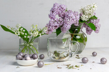 Hot herbal tea in glass, spoon, lilac and lily of the valley flowers on white background, easter egg sweets