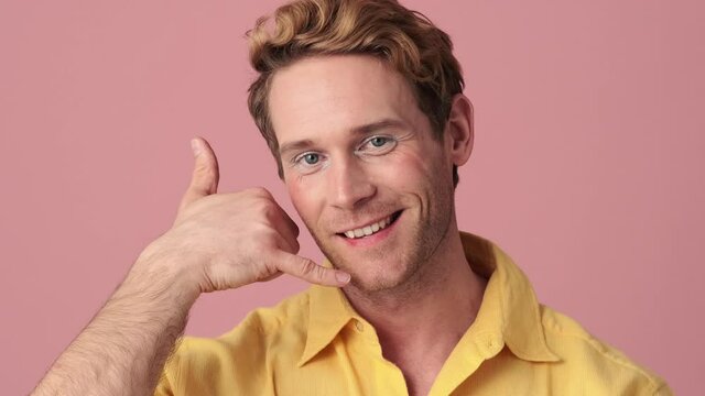 The close-up view of a happy man with makeup showing a call me gesture with his hand in a pink studio
