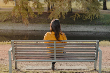 Horizontal shot of the back of a Woman in a yellow sweater sitting on a bench in a park alone looking out over a stream. Sunbeams illuminate the scene. Social distancing concept..