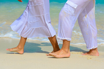 Fototapeta na wymiar Close-up of the legs of couple walking barefoot on the beach in white outfits
