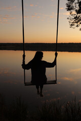 silhouette of a romantic young woman on a swing over lake at sunset. Young girl traveler sitting on the swing in beautiful nature, view on the lake