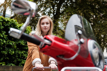 Obraz premium Young blonde woman posing behind of a red scooter.