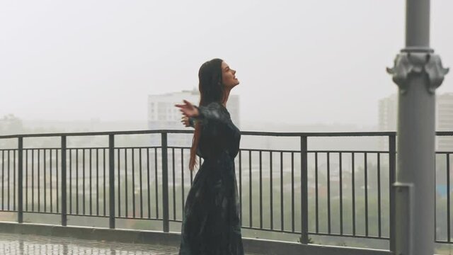 Happy pretty woman in long wet chiffon dress turns dancing under heavy rain on viewing deck against foggy city on summer day slow motion
