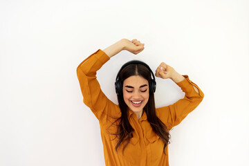 Fototapeta na wymiar Beautiful brunette woman in ocher shirt and black headphones listens to music and smiling on white background. Pretty girl dancing and singing in studio, playing music on her headphones and smiling.