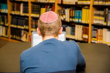 Young man wearing a yarmulke from the back doing work on a laptop in a library with colorful books...
