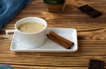 a cup of latte with froth stands on a square saucer on a wooden table. Cinnamon sticks, dark chocolate and coffee pieces. High quality photo