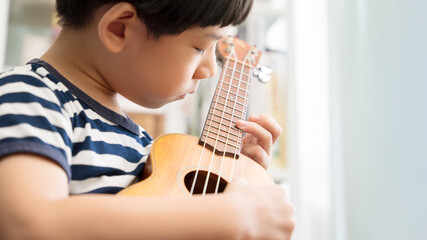 Fototapeta premium Cute Asian little boy playing and practicing ukulele skills with focus and concentration at home. Acoustic Music instrument, Child brain development activity, Homeschool and music therapy concept.