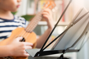 Online music class lesson concept. Cute Asian little boy playing and practicing ukulele skills with focus and concentration via computer tablet at home. Child brain development activity, Homeschool.