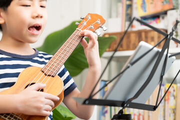 Fototapeta premium Online music class lesson concept. Cute Asian little boy playing and practicing ukulele skills with fun and happiness via computer tablet at home. Child brain development activity, Homeschool.