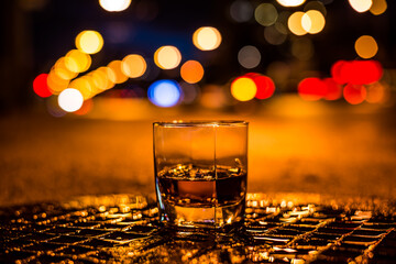 Lights of the city at night through the glass of alcohol, the night avenue and sidewalk. View from the glass level with brandy standing on the wet manhole