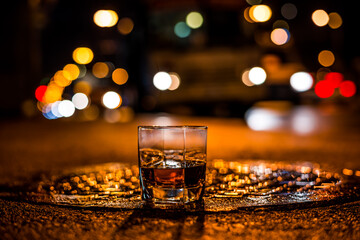 Lights of the city at night through the glass of alcohol, the night avenue with driving bus. View from the glass level with brandy standing on the wet manhole
