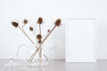 Empty white frame mockup. Glass teapot and dried thistle flowers on light table.