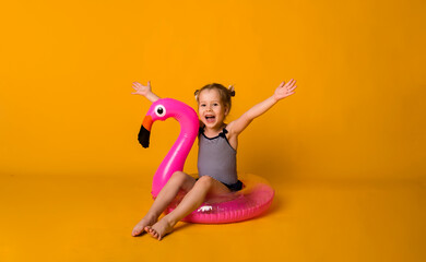 happy little blonde girl in a striped swimsuit sits in a pink inflatable circle with a flamingo and looks at the camera on a yellow background with a place for text. Summer holidays with a child