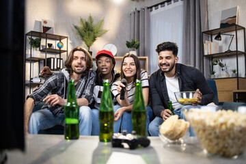 Carefree diverse friends with beer and snacks looking at TV screen while singing in microphone. For young people spending leisure time at home with karaoke.