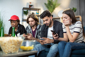 Four multi ethnic friends sitting together on couch and using personal smartphones. Home party with modern gadgets. Addiction from social networks.