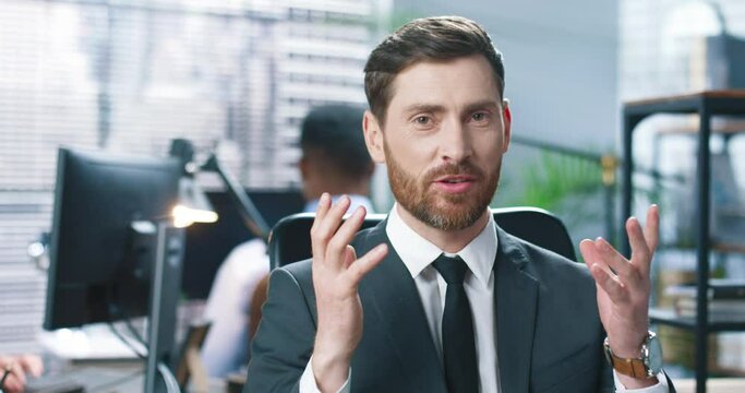Close up portrait of Caucasian young handsome businessman in suit sitting at workplace in cabinet and speaking to camera, recruitment, business trainer, man employee working at office