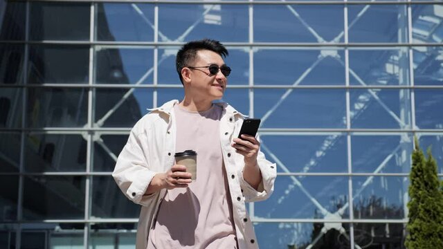 Successful happy Asian Businessman in Casual Clothes is texting Messages on modern Smartphone. Young Man is walking in Business Quartal, having good Mood, looking Satisfied. Successful Lifestyle. Men.