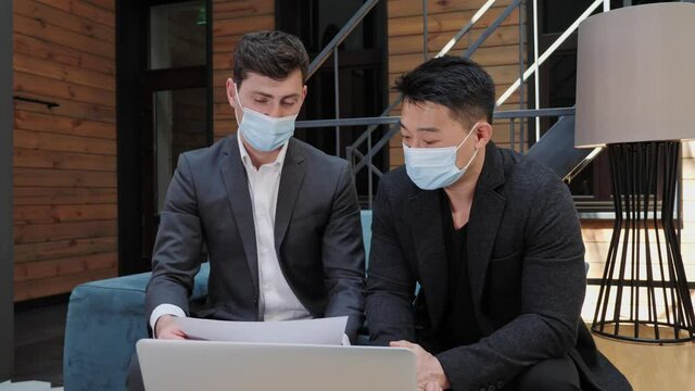 Two diverse multinational businessman in medical mask holding documents and discussing partnership details in front of the laptop in the meeting room International collaboration concept during covid19