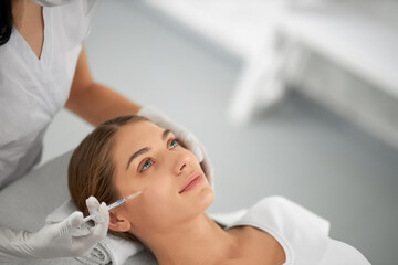 Side view portrait of smiling beautiful young woman in white shirt doing procedure for improvements face skin. Concept of special cosmetics injection for anti-aging in beauty salon.