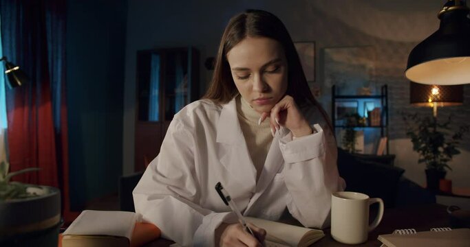 Young woman wearing medical uniform gown, female intern working at night with a computer in a medical office. Doctor nurse using laptop and video call, writing notes, overtime at the workplace.