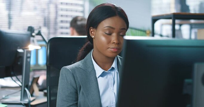 Close up of young African American pretty joyful woman office manager in cabinet room sitting at workplace surfing internet on computer, looking at camera and smiling, businesswoman concept