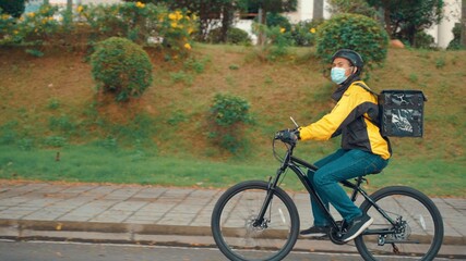 Obraz premium Food Delivery Man Wearing Protective Medical Face Mask and Thermal Backpack Rides a Bike to Deliver Order. Young Asian man working as a food delivery person. He riding a bike for sustainable way.