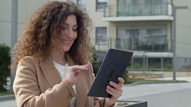 Curly-haired woman middle-aged walks along a deserted city street. She communicates with someone through a tablet.