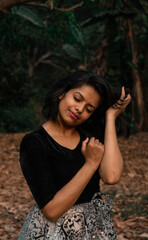 Portrait of a thoughtful young girl smiling with eyes closed and her head resting on her hands wearing a black dress posing in a dark grungy forest