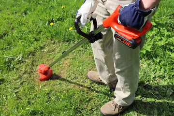 Man holds an electric trimmer in his hands and mow grass in meadow, on sunny day with selective focus