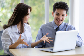 Beautiful black long hair Asian businesswoman and gook looking cute Asian businessman sitting together and watching the screen of laptop notebook computer in office with smile face