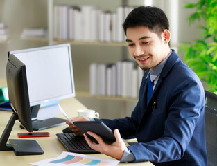 Successful young Asian businessman smiles happily while seeing good news on tablet in office. Business and success concept.