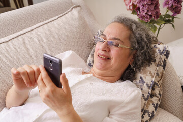 Elderly woman is surfing the internet with her phone at her home. Housewife ordering grocery shopping from her home with her smart phone. Elderly woman using a smart phone. Old woman shopping online.