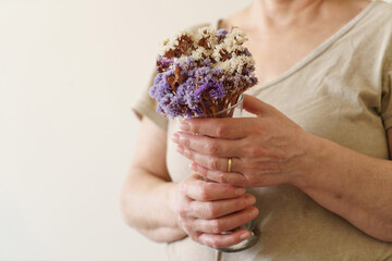 the old woman with only her hands visible is holding a vase with dried flowers. Concept photo showing dried flowers and old woman's hands together.