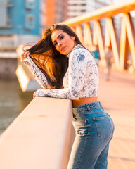 A brunette Latin girl in jeans in the city at sunset. On a bridge looking at the city river, vertical photo