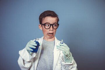cool school boy with thick black glasses is dressed as scientist with white coat and experimenting with blue liquids in front of blue background