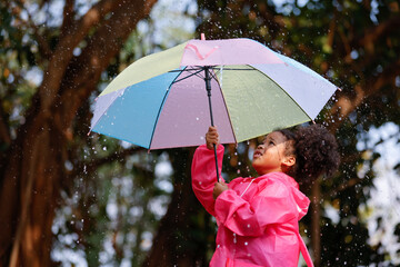 Happy funny child curly black hair with umbrella under the autumn shower. Girl is wearing pink raincoat and enjoying rainfall. Kid playing on the nature outdoors.