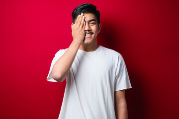 Young handsome asian man wearing white tshirt standing covering one eye with hand, confident smile on face and surprise emotion.