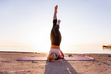 Young blonde woman in sportswear performs yoga asanas, exercises on the seashore at sunrise. The...