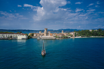 Fototapeta na wymiar The Old Sailing Ship is sailing near Sirmione Castle. Rocca Scaligera Castle in Sirmione Lake Garda Italy. Clouds in the blue sky. Aerial view.