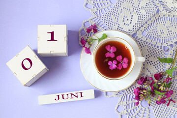 Calendar for June 1: cubes with the numbers 0 and 1, the name of the month of June in English, a cup of tea with purple flowers in it, a gray openwork napkin on a light background, top view