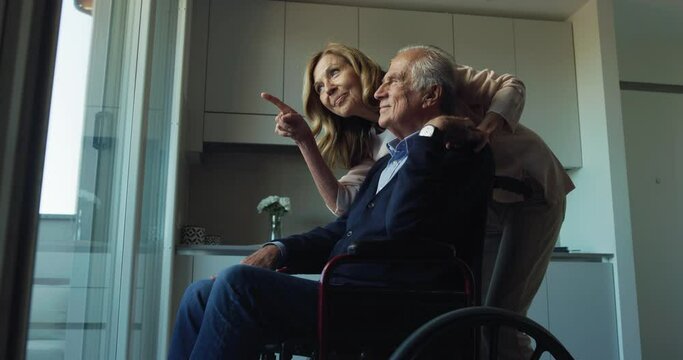 Cinematic shot of smiling mature wife hugging disable husband in wheelchair as sign of forever timeless love while looking through window at home. Concept of life, family, marriage, happiness, age.
