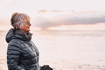 Senior woman in outdoor excursion at sea looking at sunset. Standing on cliff, horizon over water