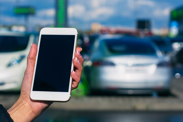 Girl holds a smartphone in his hands. Mock up phone with white screen against the background of an electric car at a charging station.