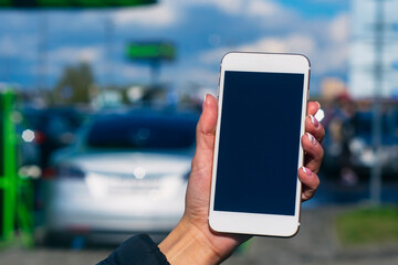 Girl holds a smartphone in his hands. Mock up phone with white screen against the background of an electric car at a charging station.